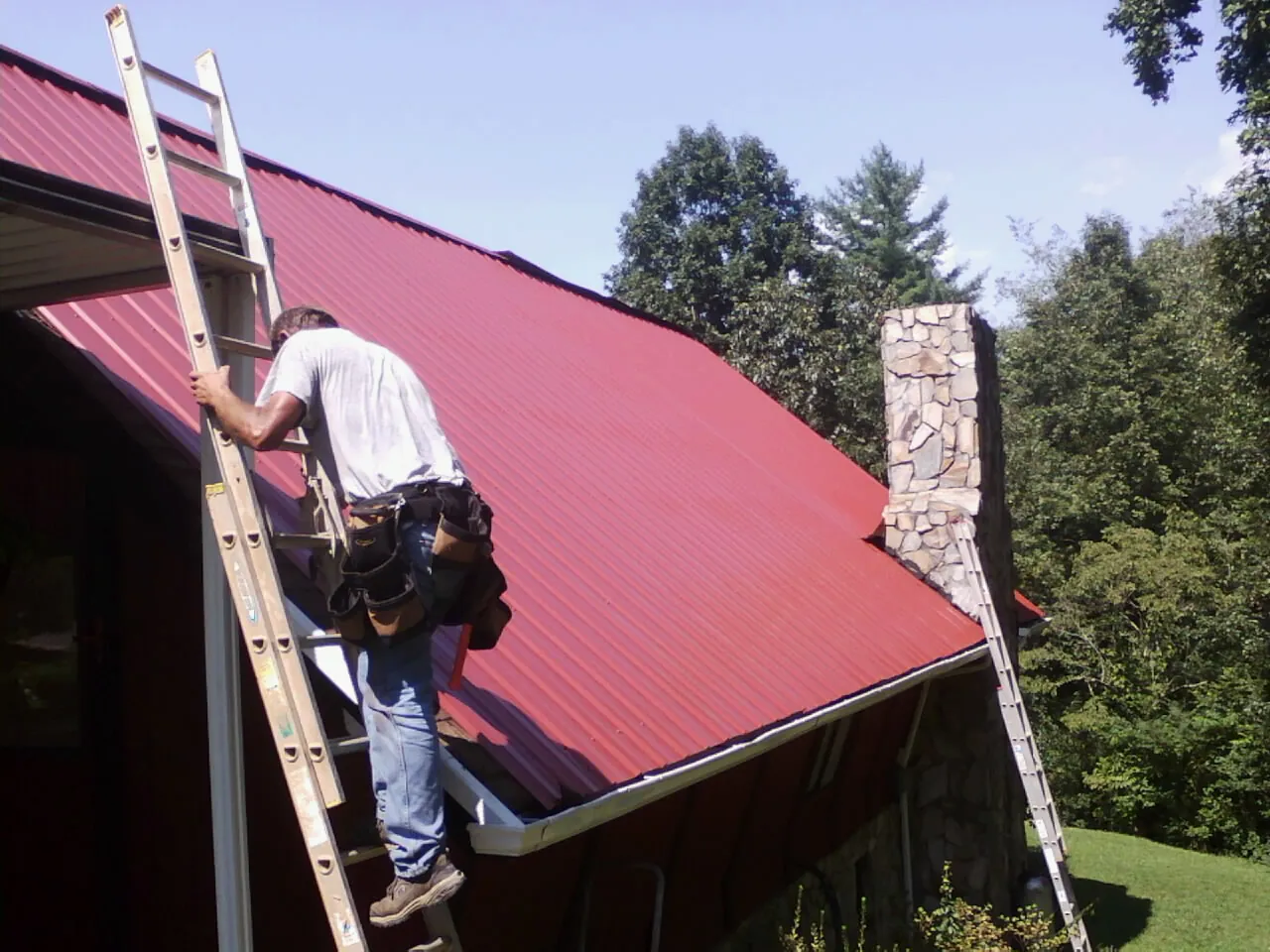Roofer on a ladder beside a red metal roof and stone chimney — Soffit & Fascia Repair by Roofing Brothers in Pine Grove